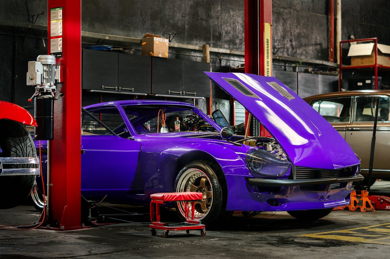 A classic car with a vibrant purple color in a mechanical workshop with its hood open.