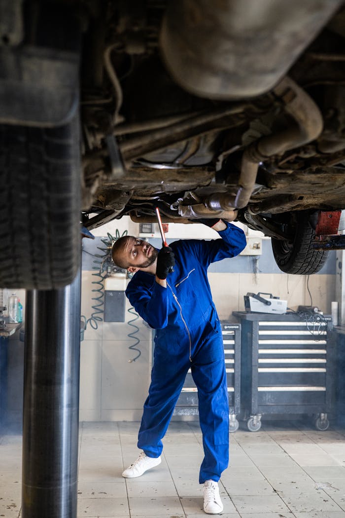 A female mechanic in blue coveralls inspects a car's undercarriage in an auto repair shop.